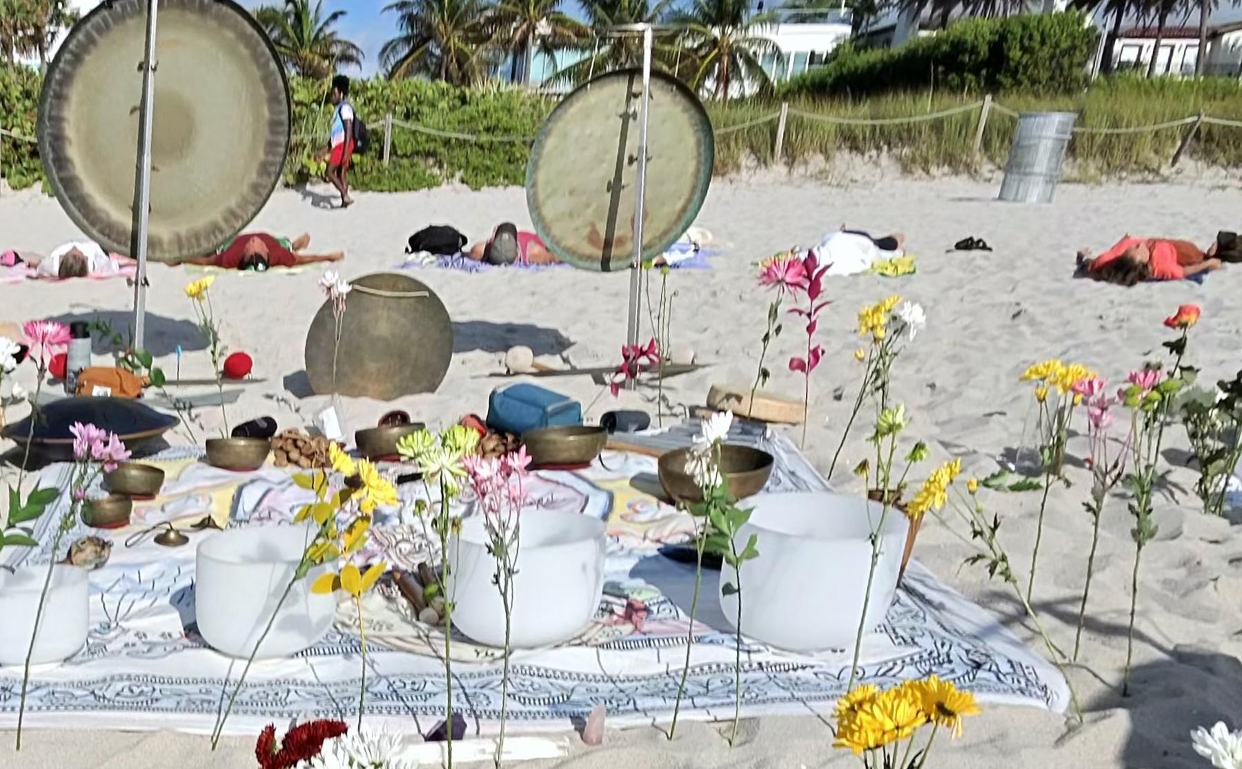 Two gongs and an altar of bowls and flowers on the sand, with guests resting behind